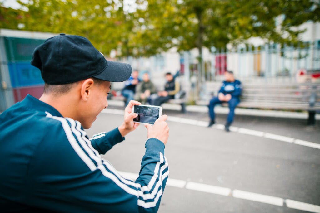 Photo from behind showing a teenage boy taking a photo on a smartphone of his friends who are sitting on benches in a public space