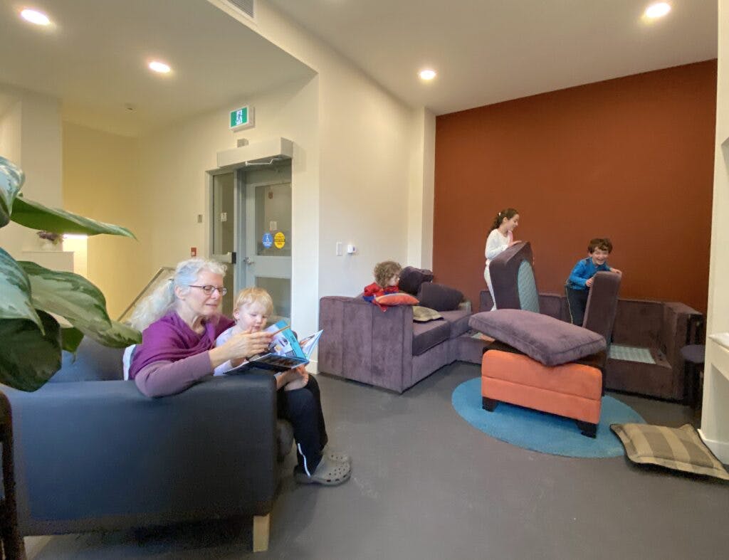 Photo of residents hanging out in the little mountain cohousing lobby. An older woman reads a story to a young kid on an airmchair. in the background, three kids play with couch cushions