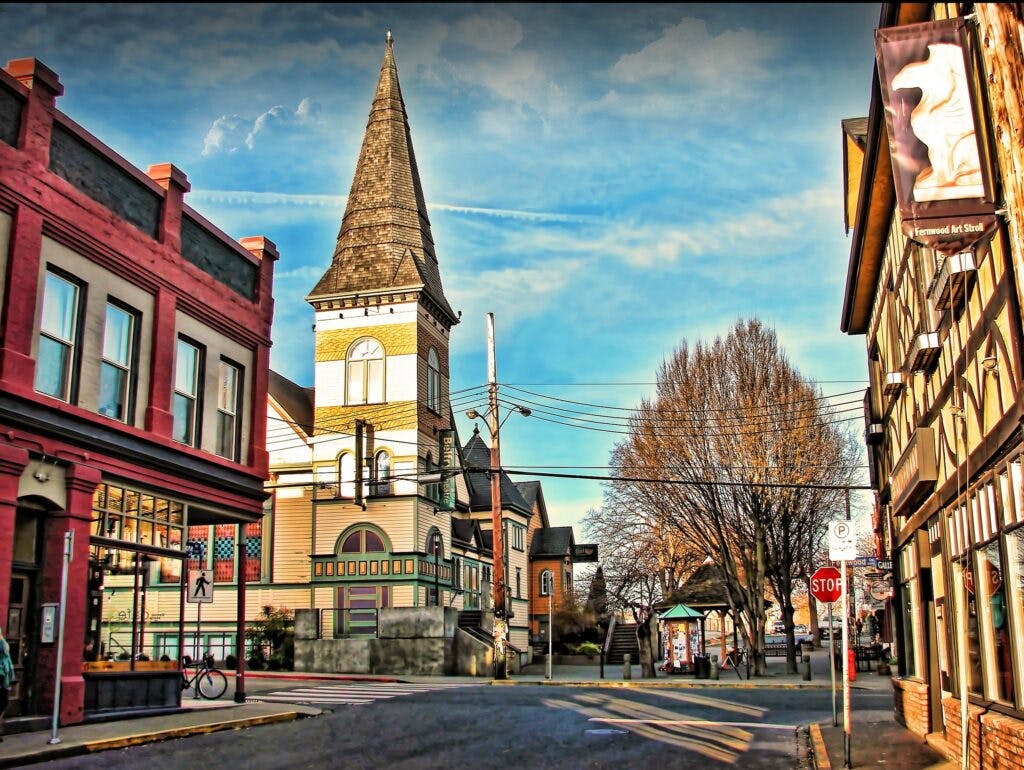 Photo of the main street in Fernwood, Victoria, BC. Quiet streets are lined with local shops painted in different colours. At the end of the street there is a building with a steeple, and a small public area with benches
