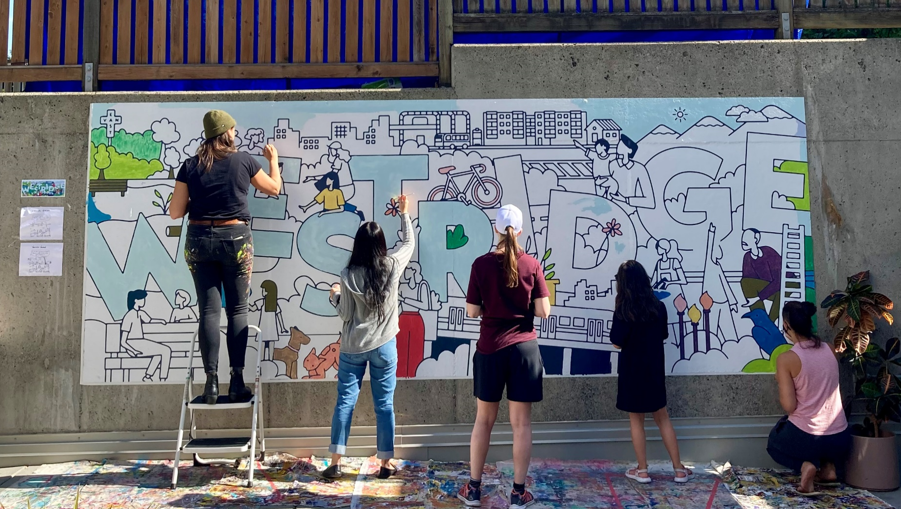 Photo of 6 people standing/squatting side by side and colouring in a large horizontal mural on a wall. The mural says "Westridge" in large block letters, and includes illustrations of people, buildings, flowers, bikes and more.