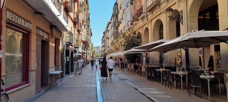 Photo of a pedestrian street in Logrono, Spain. The street is straight and narrow, lined with 3-4 storey buildings with restaurants and shops at ground level and housing with balconies above. People walk along the street. On the right side, there is a patio with umbrellas.