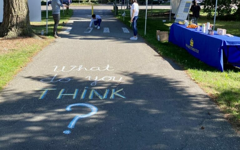 Photo of a pop-up engagement event in a public park. Chalk letters on the ground read, "What do you think?" There is a table set up with free food and snacks, and several tents to provide shade